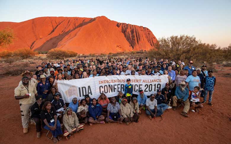 A large number of participants at the Indigenous Desert Alliance Conference holding a banner showing the conference name with Uluru in the background