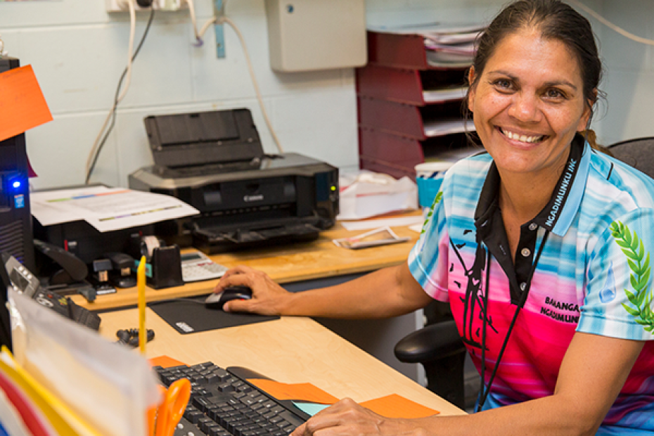 A woman in a bright coloured t-shirt sits at a desk in an office. She has dark hair and is typing on a computer, and smiling.