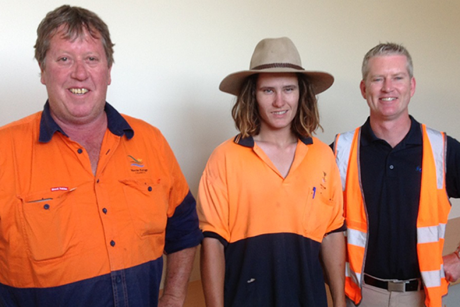 Three men stand wearing predominantly orange work wear. The one in the middle is younger and wears a broad brimmed hat.