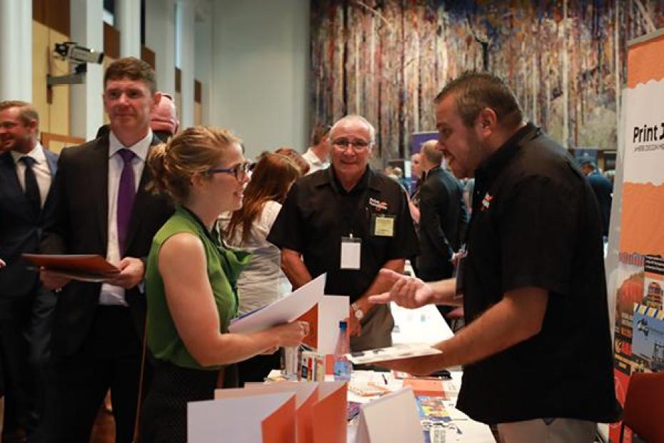 A young women listens intently to an Indigenous business man who is speaking about his print products at a trade fair. Many people are moving around them.